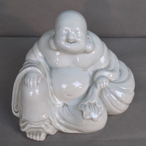 May include: White porcelain figurine of a smiling, seated Buddha. The Buddha is wearing a robe and has a large belly.