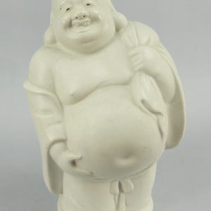 May include: White ceramic figurine of a smiling, chubby Buddha holding a bag over his shoulder.