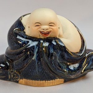 May include: A ceramic figurine of a laughing Buddha, sitting with his hands clasped in his lap. He is wearing a blue and white robe with gold accents.