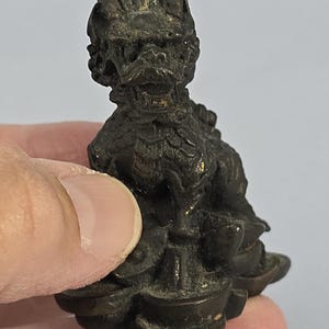 May include: A small, dark bronze-colored figurine of a mythical creature, possibly a dragon or lion. The detailed sculpture features a textured surface and is held in a person's hand. The figurine is resting on a decorative base.
