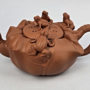 May include: A terracotta teapot with a lotus design and a lid with five frogs on top. The teapot has a brown glaze and a curved spout.
