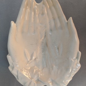 May include: White ceramic dish shaped like two hands clasped together in prayer. The dish has a smooth, glossy finish and a slightly textured surface.