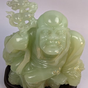 May include: A pale green jade sculpture of a seated figure holding a staff with a dragon-like design. The figure has a serene expression and is mounted on a dark wooden base. The sculpture is intricately carved and detailed.