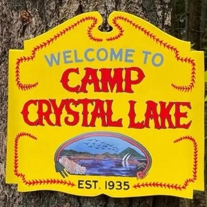 May include: A yellow sign with a decorative border resembling baseball stitching. The text reads WELCOME TO CAMP CRYSTAL LAKE in red and blue, with a central illustration of a lake, mountains, and birds, and the text EST. 1935 at the bottom.