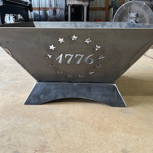 Puede incluir: Un pozo de fuego de metal con un diseño en forma de estrella y el número 1776 recortado. El pozo de fuego tiene una base cuadrada con un borde redondeado.