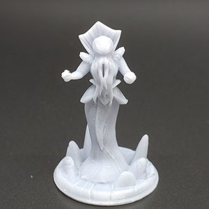 May include: A light gray figurine of a humanoid figure with a crown-like headpiece and tentacles. The figure is standing on a circular base with stylized wave-like shapes. The figurine is made of a solid, smooth material.