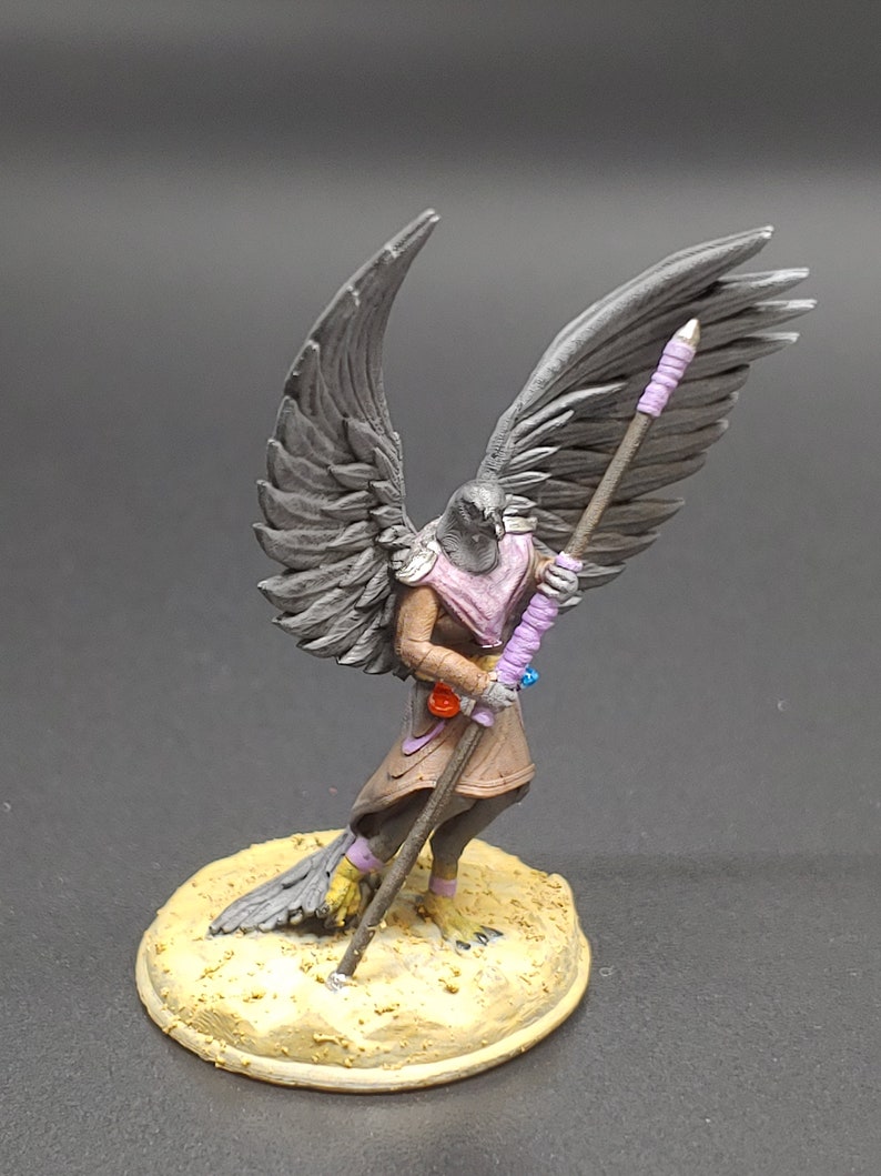 May include: A miniature figurine of a bird-like humanoid with outstretched wings. The figure is predominantly grey, with a pink scarf and brown clothing. It holds a staff wrapped in purple. The base is a tan colour.