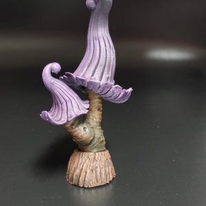 May include: A purple and brown miniature mushroom sculpture with a whimsical design. The mushroom has a tall, slender stem and a wide, flat cap with a curled edge. The cap is painted in a light purple color, while the stem is painted in a brown color.