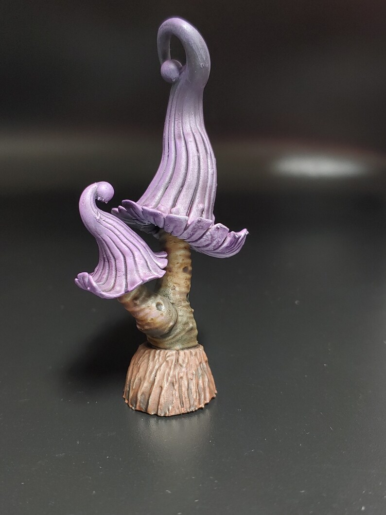 May include: A purple and brown miniature mushroom sculpture with a whimsical design. The mushroom has a tall, slender stem and a wide, flat cap with a curled edge. The cap is painted in a light purple color, while the stem is painted in a brown color.
