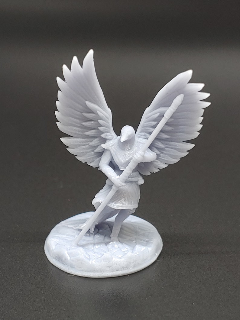 May include: A light grey figurine of an angel with large, detailed wings, holding a staff. The angel stands on a circular base with a textured surface. The figure is a detailed representation of a mythological or fantasy character.