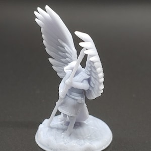 May include: A light grey miniature figurine of an angel with large, detailed wings. The angel is holding a staff and stands on a textured, circular base. The figurine is made of a solid, light grey material.