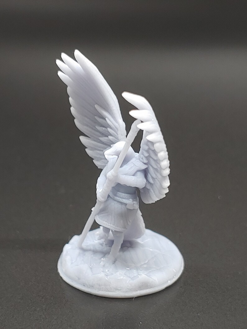 May include: A light grey miniature figurine of an angel with large, detailed wings. The angel is holding a staff and stands on a textured, circular base. The figurine is made of a solid, light grey material.