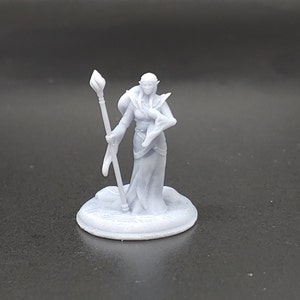 May include: A white 3D printed miniature of an elf wizard holding a staff. The elf is wearing a long robe and has a pointed hat. The miniature is standing on a round base.