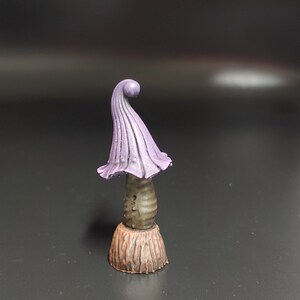 May include: A small purple and brown mushroom figurine with a curled top. The mushroom is painted with a light brown base and a darker brown stem. The top of the mushroom is a vibrant purple color.