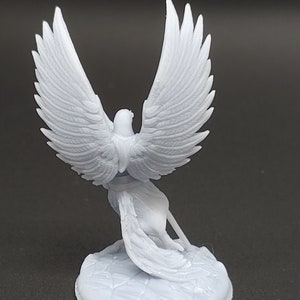 May include: A light grey figurine of a bird with large, detailed wings, perched on a textured base. The bird is sculpted in a stylized pose, with its wings spread wide. The figurine is made of a smooth material.