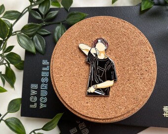 May include: A metal pin featuring a stylized illustration of a person in a black shirt holding a microphone. The pin is set on a cork coaster, with a black card that reads "LOVE YOURSELF" in white text. Green leaves surround the objects.