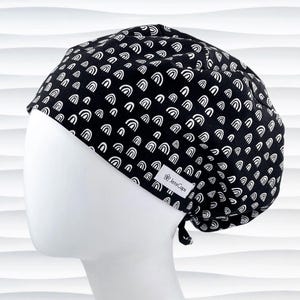 May include: Black surgical cap with a repeating white rainbow pattern. The cap is tied at the back and features a small white tag with the text "JensCaps". The cap is displayed on a white mannequin head.