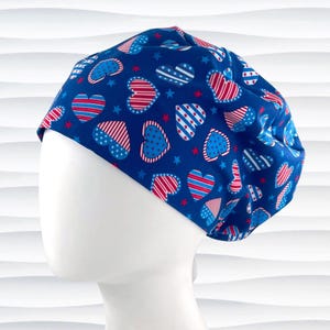 May include: A blue scrub cap with a pattern of red and white striped hearts, blue hearts with white stars, and red stars. The cap is on a white mannequin head.