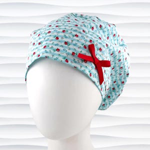May include: A scrub cap featuring a light blue and white striped pattern with a small red floral print throughout. The cap is accented with a bright red ribbon bow attached to the side, providing a decorative detail to the fabric head covering.