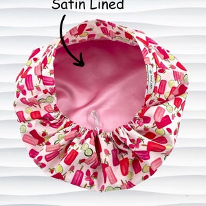 May include: A satin-lined bonnet with a pink interior and a white exterior patterned with red popsicles, strawberries, and lime slices. The text "Satin Lined" is above the bonnet.