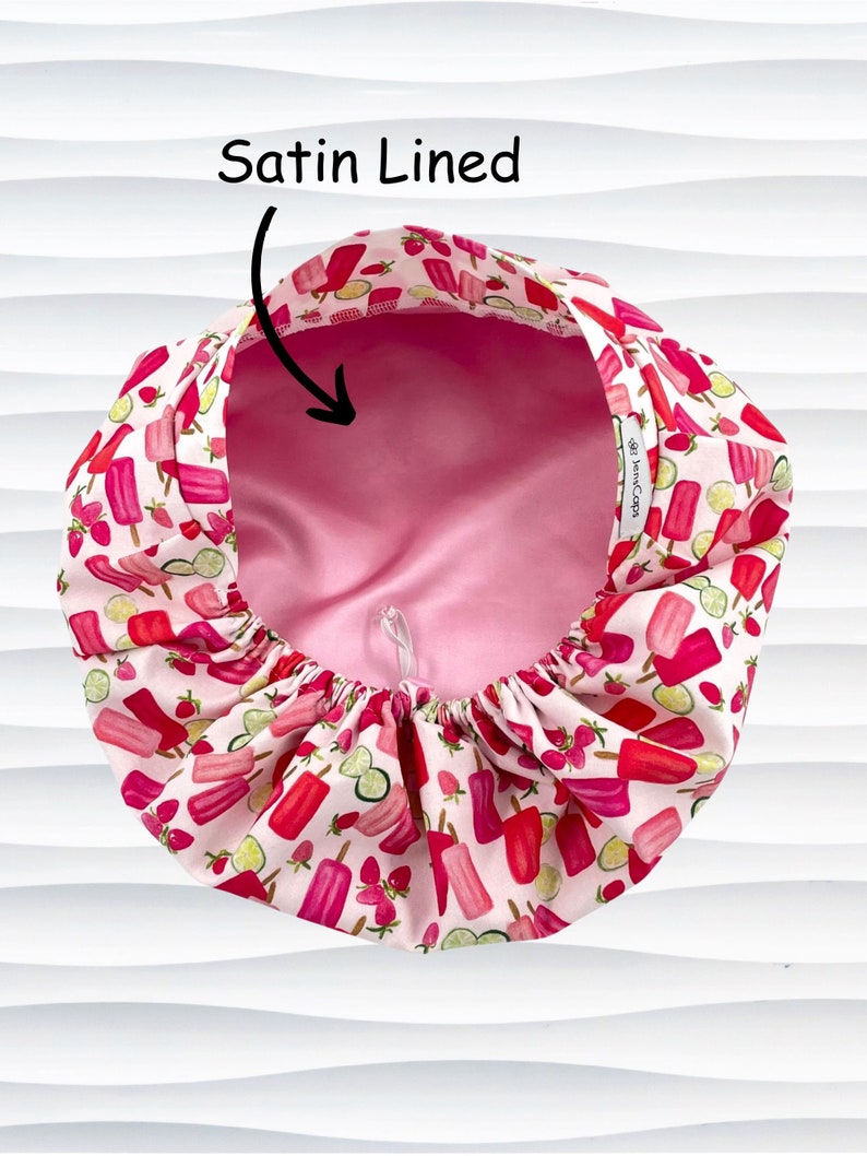 May include: A satin-lined bonnet with a pink interior and a white exterior patterned with red popsicles, strawberries, and lime slices. The text "Satin Lined" is above the bonnet.
