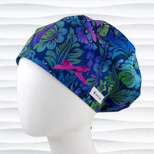 May include: A scrub cap featuring a dark blue background with a vibrant floral and leaf pattern in shades of purple, light blue, and green. The cap includes a small magenta ribbon bow on the side and a white label with the text JensCaps.