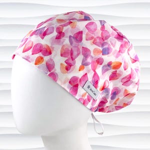 May include: A white scrub cap with a pink and purple watercolor floral pattern. The cap has a tie at the back and a small logo that says "JemsCaps".