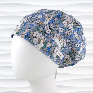 May include: A blue floral scrub cap featuring white rabbits and flowers. The cap has a tag that reads "JensCaps". The fabric is a mix of blue, white, and yellow.