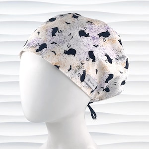 May include: A scrub cap featuring a white background with a pattern of black cats and small bats. The fabric has a mottled texture in shades of purple and orange. A small rectangular tag on the side displays the text JensCaps with a flower icon.