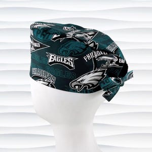 May include: A dark teal scrub cap featuring a repeating pattern of Philadelphia Eagles logos, including the eagle head emblem, the wordmark, and pennant designs. The cap has a tie-back closure and is displayed on a white mannequin head form.