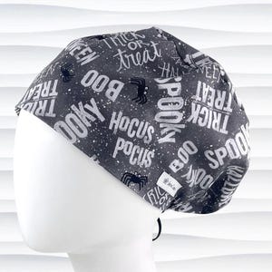 May include: A dark gray scrub cap featuring a white speckled pattern and repeating Halloween-themed text including "Trick or Treat," "Boo," "Spooky," and "Hocus Pocus," interspersed with small black spider illustrations and a small white brand tag.