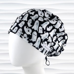 May include: Black and white patterned scrub cap featuring a repeating ghost design. The cap is made of fabric and has a tie in the back for adjustment. The brand name "JensCaps" is visible on a small white tag.