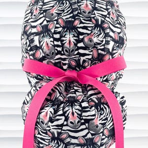 May include: A patterned scrub cap featuring a repeating zebra face design in black and white. The cap is tied with a bright pink ribbon. The zebra faces have pink noses and ears, and blue eyes.