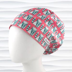 May include: A scrub cap featuring a vibrant coral pink background with a repeating pattern of stylized teal and white raccoons. The cap includes a small white fabric label on the side with the text JeniCaps printed in black lettering.