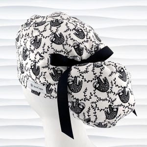 May include: A white scrub cap featuring a black sloth and vine pattern. The cap has a black bow and is designed to be worn during medical procedures. The brand name "JensCaps" is visible on the side.
