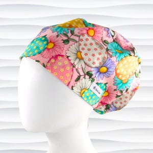 May include: Pink scrub hat with a colorful Easter egg and flower pattern. The hat has a small white label with the text "Scrub Caps" on it.