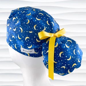 May include: A blue scrub cap with a celestial pattern of yellow crescent moons and stars. The cap has a yellow ribbon and a small white tag that reads "JensCaps".