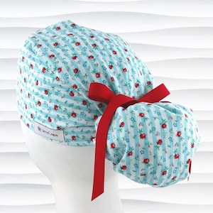 May include: A light blue and white striped scrub cap with a floral pattern of small red roses. The cap has a red ribbon tied in a bow at the back. The brand name "JensCaps" is visible on the side.