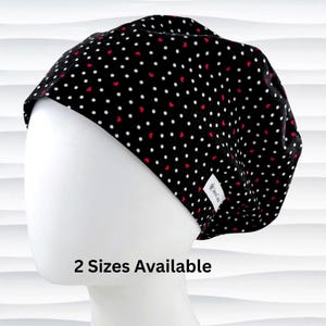 May include: A black surgical scrub cap with a pattern of white polka dots and small red hearts. The cap is displayed on a white mannequin head. The text "2 Sizes Available" is visible at the bottom.