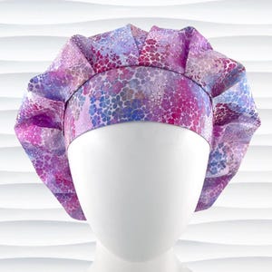 May include: A bouffant-style scrub cap featuring a vibrant floral pattern in shades of purple, pink, and blue. The cap has a wide, structured headband and a gathered, pleated crown that provides a voluminous shape for full hair coverage.