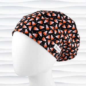 May include: A scrub cap featuring a black background with a repeating pattern of candy corn shapes in orange, white, and pink. The cap includes a small white rectangular fabric label on the side with the text JensCaps printed in black ink.