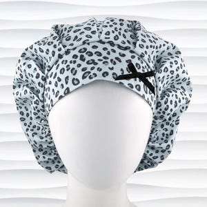 May include: A light blue scrub cap featuring a dark gray leopard print pattern. The cap has a gathered, pleated design around the crown and a small, black fabric bow attached to the front band, providing a secure and adjustable fit for the wearer.