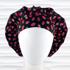 May include: A black scrub cap featuring a repeating pattern of small red strawberries with green leaves and smiling faces. The cap has a wide, structured front band and a gathered, pleated crown design that provides a full, rounded shape.
