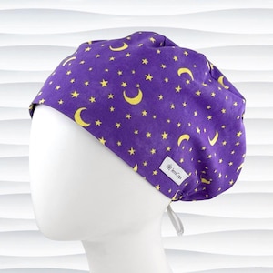 May include: A purple scrub cap featuring a repeating pattern of yellow crescent moons and stars. The cap has a rounded crown and a small white fabric tag on the side with the text JensCaps printed in black. The back includes thin fabric ties.