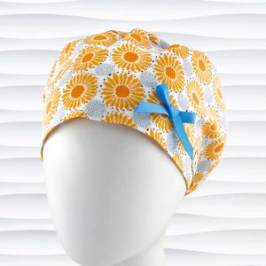 May include: A scrub cap featuring a repeating pattern of yellow sunflowers and small light blue circular clusters on a white background. The cap is accented with a bright light blue ribbon bow attached to the side, providing a decorative detail.