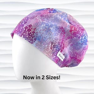 May include: A scrub cap featuring a vibrant floral pattern in shades of pink, purple, and blue. The fabric has a repeating small flower design. A small white rectangular tag with the text JensCaps is sewn onto the side. Text reads Now in 2 Sizes!