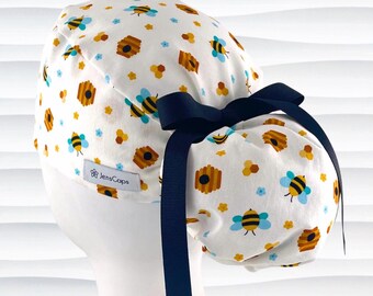 May include: White scrub cap with a repeating pattern of bees, beehives, and flowers in yellow, blue, and brown. A navy blue bow is tied on the back. The cap has the text "JensCaps" on the side.
