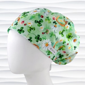 May include: A light green scrub cap with a floral pattern featuring white daisies, green clovers, ladybugs, and gold horseshoes. The cap is made of a soft fabric and has a tag that reads "JensCaps".