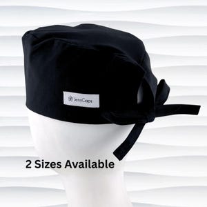 May include: A black surgical scrub cap with a tie closure, displayed on a white mannequin head. The cap has a small white label with the text "JensCaps". The text "2 Sizes Available" is displayed below the cap.