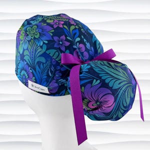 May include: A scrub cap featuring a dark blue fabric with a vibrant floral and leaf pattern in shades of purple, teal, and green. The cap includes a gathered back section secured with a purple ribbon bow and a white label reading JensCaps.